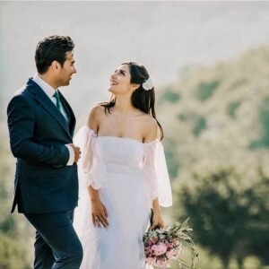 Escena romántica en la naturaleza; la novia sostiene su ramo y mira al cielo, mientras el novio en traje azul la observa con ternura.