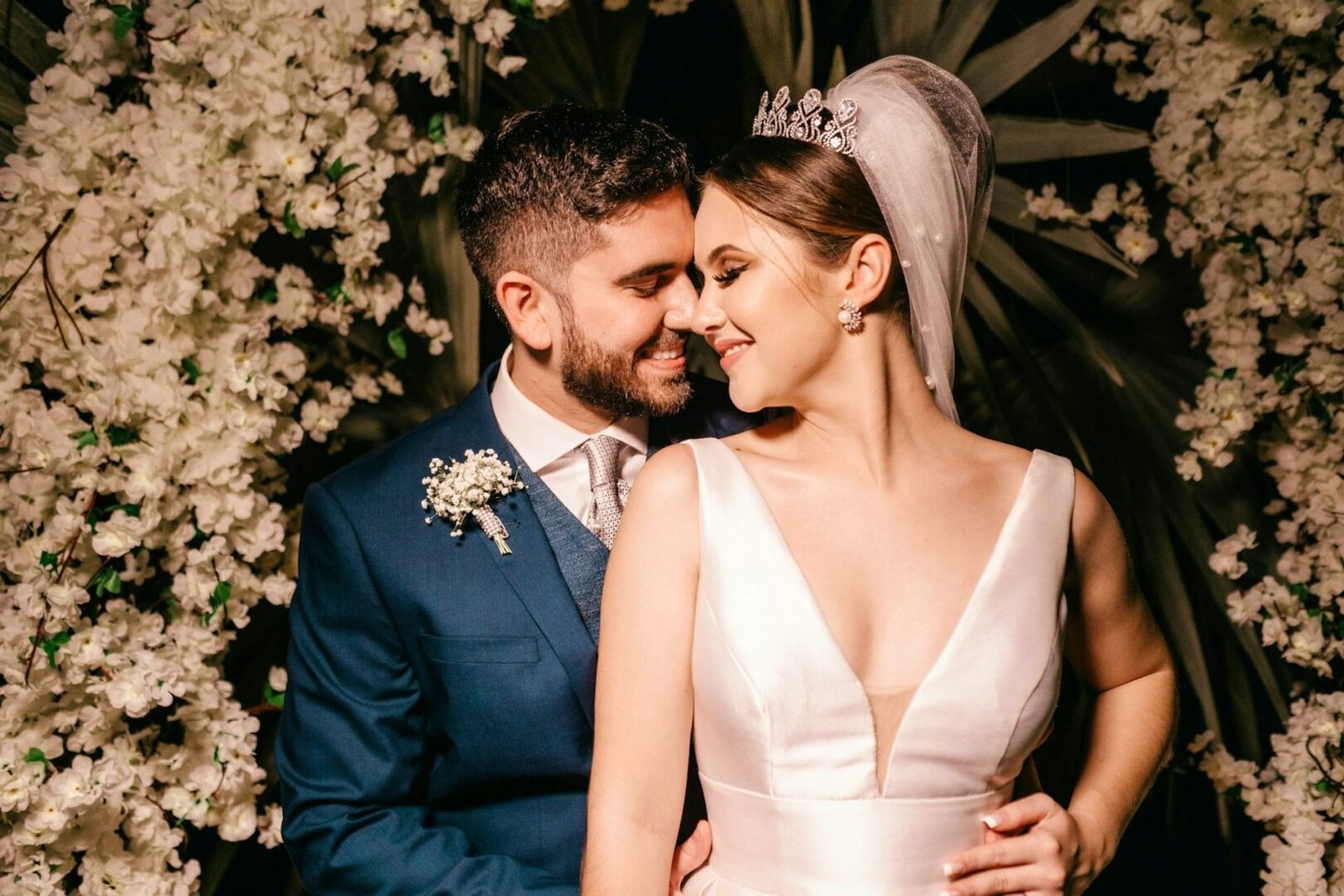 Novios el día de su boda, sonriendo abrazados de frente bajo un arco de flores blancas, a punto de besarse
